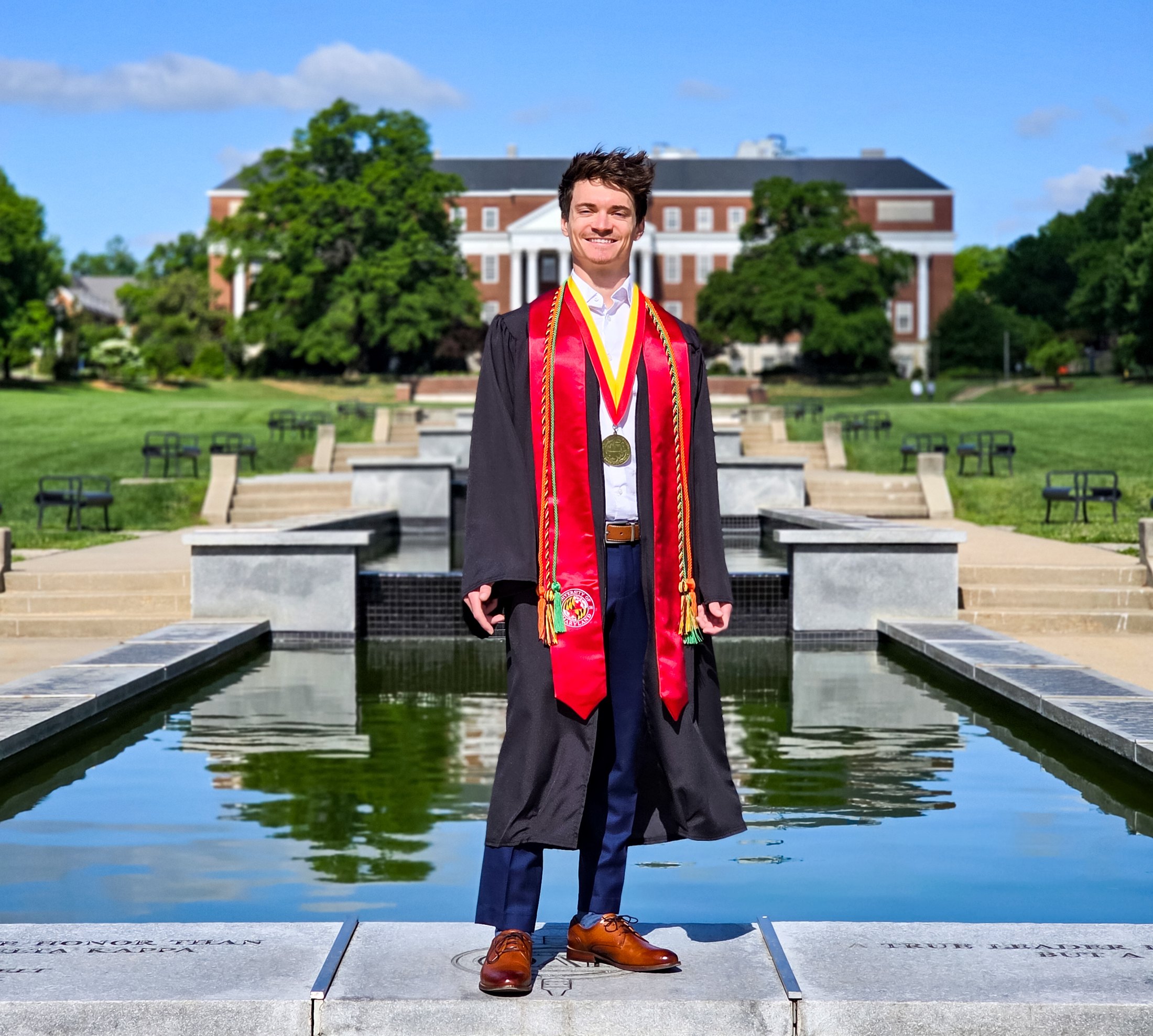 Graduate in cap-and-gown regalia with a red stole, standing at a reflecting pool on the University of Maryland campus