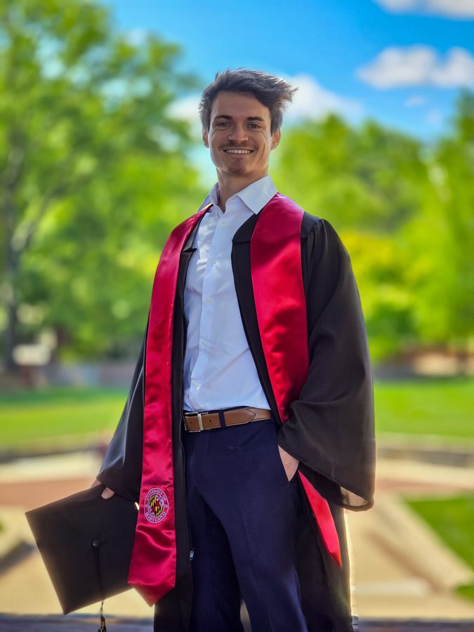 Smiling graduate holding his cap, wearing a red stole, trees softly blurred behind